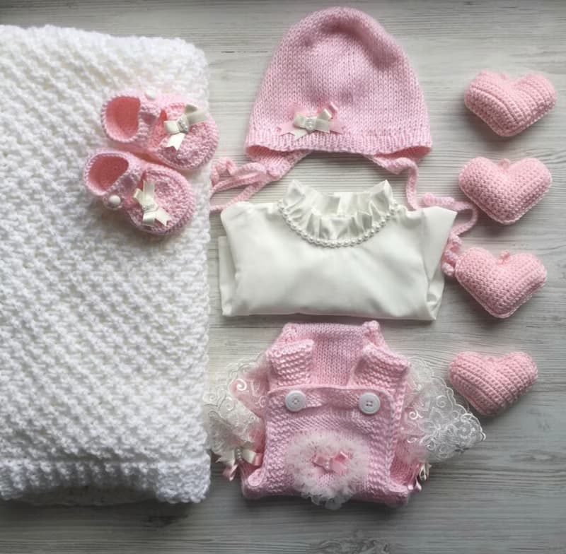 Pink knit romper, hat booties, blouse and white blanket with four pink hearts.